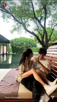 a man and woman sitting on top of a wooden bench next to a tree in front of a pool