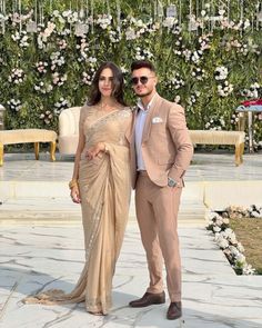 two people standing next to each other in front of a flower covered wall with white flowers