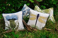 three decorative pillows with dragonflies and thistles on them sitting in the grass next to trees