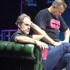 two men sitting on top of green couches in front of a stage with blue lights