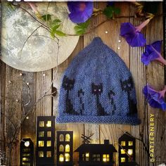 a blue knitted hat with black cats on it and purple flowers in the background