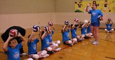 a group of young children holding up volleyball balls on top of a gym floor with their hands in the air