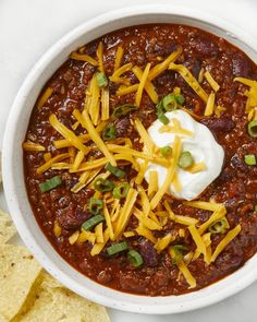 a bowl of chili with cheese and sour cream on top, next to tortilla chips