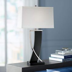 a lamp on a night stand next to a bed with books and a window in the background