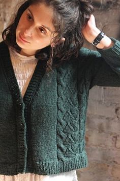 a woman wearing a green cardigan standing in front of a brick wall with her hands on her head