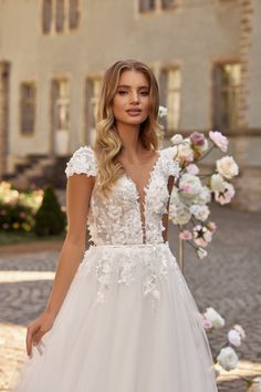 a woman in a white wedding dress standing on a cobblestone street with flowers behind her