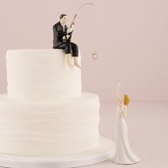 a bride and groom figurine sitting on top of a wedding cake, next to a miniature fishing rod