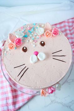 a cake decorated to look like a cat with flowers on its head is sitting on a pink and white checkered tablecloth