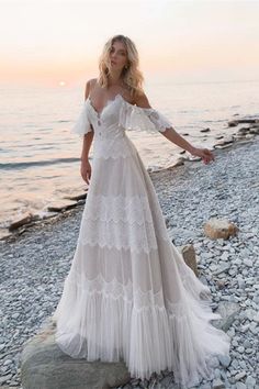 a woman standing on top of a rocky beach next to the ocean wearing a white dress