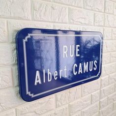 a blue street sign mounted to the side of a white brick wall with words rue albert camus on it