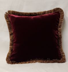 a dark red velvet pillow with fringe trimmings on the bottom and sides, against a white background