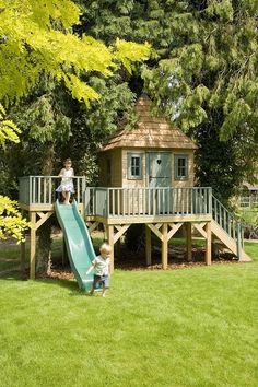 two children playing on a slide in the grass near a tree house with a deck