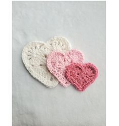 three crocheted hearts are sitting on a white surface, one is pink and the other is white