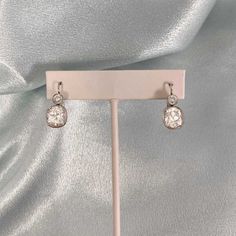 a pair of diamond earrings sitting on top of a white stand with a silver cloth behind it