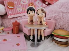 two dolls sitting at a table in front of a pink teddy bear and other toys