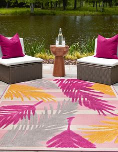 two wicker chairs with pink and yellow pillows sit on a rug in front of a lake