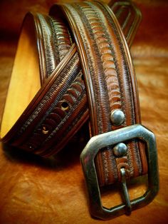 "This Brown leather belt starts as a 1-3/4\" wide piece of 9 oz. natural veg-tan leather. It is hand tooled with a Unique Matara border pattern, made using modified leather stamps, resulting in a highly textured, deep design! It is dyed a custom walnut brown and the edges are beveled, slicked smooth and rubbed with special waxes and achieve a beautiful vintage finish. The unique centerbar buckle is hand-distressed and can be worn with your own buckle as the belt uses 2 hand distressed snaps to a Artisan Brown Belt With Antique Buckle, Brown Artisan Belt With Antique Buckle, Adjustable Engraved Brown Belt, Traditional Brown Belt Buckles With Matching Belt, Traditional Brown Belt Buckles, Vintage Engraved Leather Belt, Custom Engraved Leather Belt Buckles, Traditional Leather Belt With Concho, Vintage Hand Tooled Leather Belt Buckles