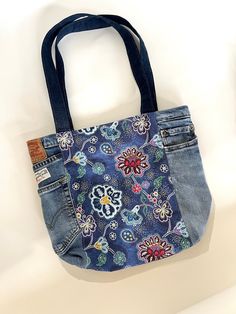 a denim bag with an embroidered flower design on the front and side pocket, hanging from a hook