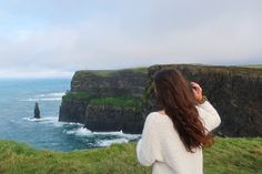 a woman standing on top of a lush green hillside next to the ocean with cliffs in the background