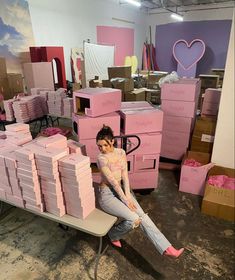 a woman sitting on top of a pile of pink boxes in a room filled with cardboard boxes