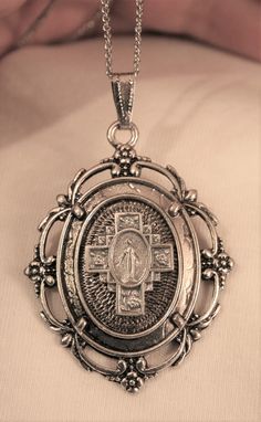 This silvertone necklace features a lovely mid-sized pendant with a scalloped outer rim with leafy accents.  The center shines with a nicely raised and sculpted cross motif with floral accents and the image of The Miraculous Mary in the center. It has a wonderful charm and character and measures 1 5/8 inches wide and 2 1/2 inches long.  The reverse has a textured finish and it is in new condition.  The chain is 20 inches and closes with a safety catch. Silver Miraculous Medal Cross Pendant Jewelry, Silver Cross Pendant With Miraculous Medal, Antique Silver Medallion With Miraculous Medal, Vintage Silver Necklace With Miraculous Medal, Vintage Silver Jewelry With Miraculous Medal, Silver Cross Locket Necklace, Silver Cross Pendant Necklace With Locket, Our Lady Of Grace, Our Lady