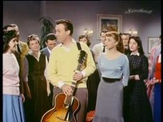 a man standing next to a woman holding a guitar in front of a group of people