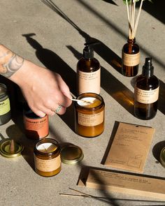 a person holding a spoon in front of some jars and candles on the ground with other items around them