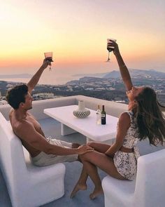 a man and woman toasting with wine glasses in front of the sun set on top of a mountain