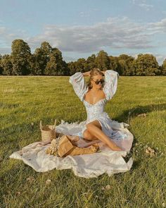 a woman sitting on top of a field next to a picnic blanket and holding her hair