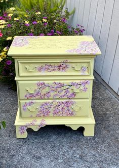 a yellow dresser with pink flowers painted on the top and bottom, sitting in front of a flower garden