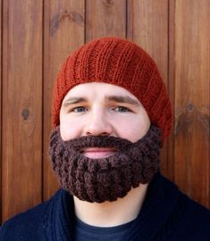 a man with a beard wearing a red knitted hat and blue sweater standing in front of a wooden wall