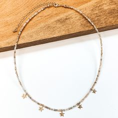 This is a beaded necklace with gold bead spacerss and five gold star charms. This neckalce includes a nude mix of beads. This necklace is pictured laying partially on a wood block on a white background. Trendy Adjustable Star Necklaces, Trendy Adjustable Star Charm Necklace, Trendy Adjustable Star-shaped Necklace, Adjustable Trendy Star Charm Necklace, Adjustable Star Shaped Charm Necklace With Adjustable Chain, Gold Star Earrings, Giddy Up Glamour, Gold Star, Star Charms