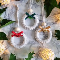 three christmas ornaments hanging from strings on a marble table with holly leaves and lights in the background