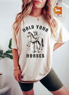 Hold Your Horses!  This funny horse shirt will look perfect on you, or buy it as a gift for the American Cowgirl in your life.  Giddy up! This Comfort Colors garment-dyed t-shirt is made with 100% ring-spun US cotton that is ethically grown and harvested. The soft-washed, garment-dyed fabric brings extra coziness to your wardrobe while the relaxed fit makes it an excellent daily choice. The double-needle stitching throughout the tee makes it highly durable while the lack of side-seams helps the shirt retain its tubular shape.  The pre-shrunk fabric ensures a consistently great fit.   Small.   ✅Chest:  18.25"  Length:  26.62" Medium✅Chest:  20.25"  Length:  28" Large.   ✅Chest:  22"       Length:  29.37" XLarge  ✅Chest:  24"       Length:  30.75" 2XLarge✅Chest:  26"       Length:  31.62" 3X Short Sleeve Cotton Top With Horse Design, Cotton Short Sleeve Top With Horse Design, Casual Cotton T-shirt With Horse Design, Cowgirl Sayings, American Cowgirl, Hold Your Horses, Oddly Specific, Horse Shirt, Funny Horse