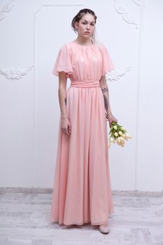 a woman in a long pink dress holding flowers and looking at the camera while standing against a white wall