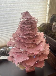 a small pink christmas tree made out of old newspaper strips and tinsel, sitting on a table in front of a window