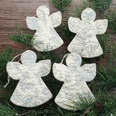 three white angel ornaments hanging from a pine branch on a wooden table with evergreen needles