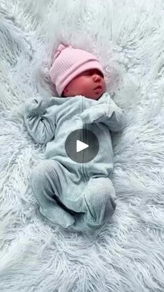 a baby laying on top of a white blanket wearing a pink hat and blue outfit