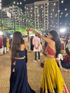 two women are dancing in the middle of a crowd at an outdoor event with string lights strung above them