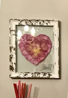 a white frame with a pink flower in the shape of a heart hanging on a wall