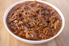 a white bowl filled with meat and sauce on top of a wooden table next to a fork