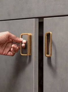 a person's hand is opening the door to a gray cabinet with brass handles