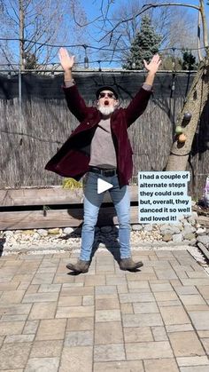 a man standing on top of a brick walkway next to a tree and fence with his hands in the air