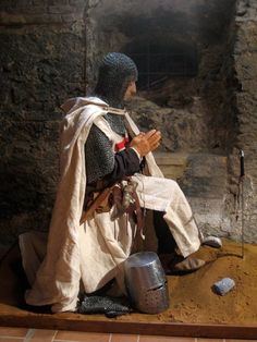 a statue of a man dressed as a knight sitting on the ground in front of a stone wall