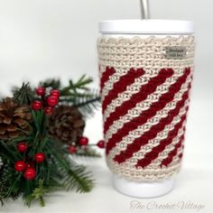 a knitted coffee cup cozying next to pine cones and evergreen needles, on a white background