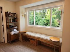 a room with a bench, bookshelf and window in it's corner