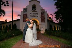 a bride and groom kissing in front of the bowers museum at sunset with palm trees