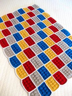 a crocheted blanket is laying on top of a white bed with red, yellow and blue squares