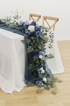 an arrangement of blue flowers and greenery on a table with two chairs in the background