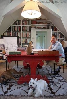 a man sitting at a red table with his dog in front of him and bookshelves behind him